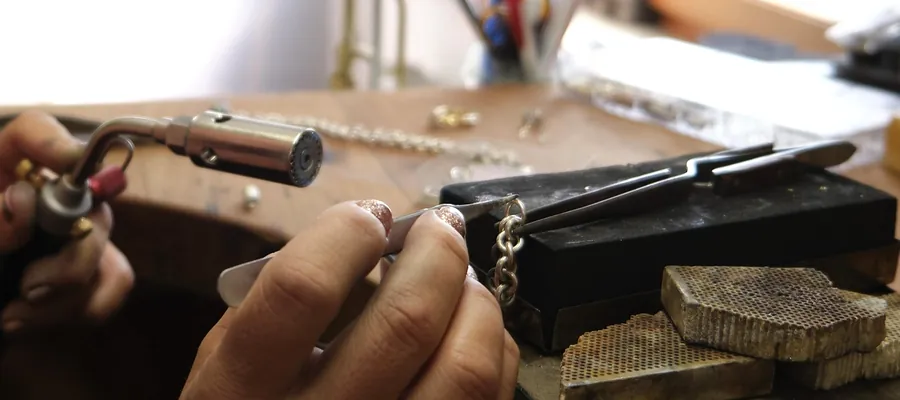 A jeweler holding a silver chain link in tweezers at a workbench, with a soldering torch and a finished chain alongside